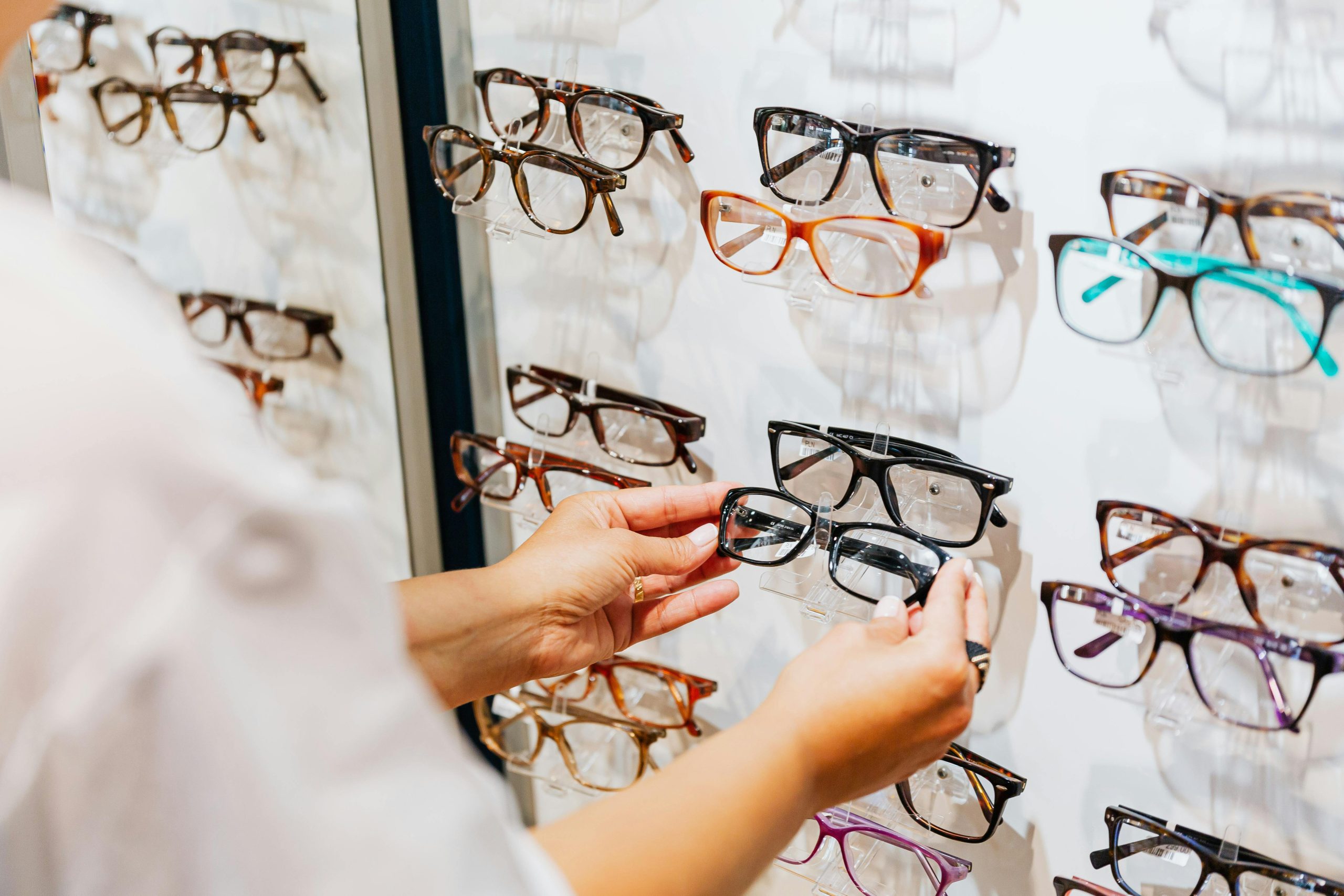 Hands selecting glasses from a display wall in an eyewear store.